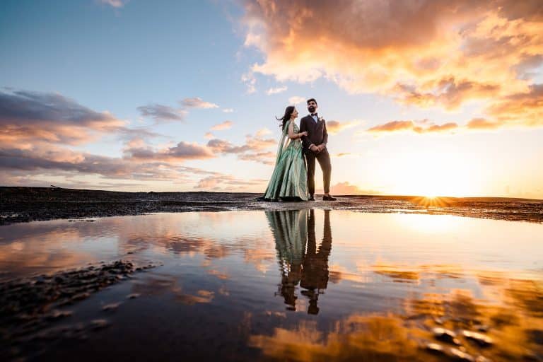 A formally dressed couple stands on a beach at sunset, the woman in a green gown and the man in a dark suit, holding hands with a reflective pool at their feet.